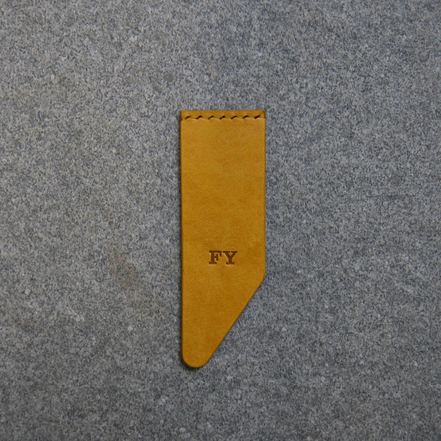 A Yellow vegetable tanned leather bookmark with the initials 'FY' embossed on it, placed on a grey granite background.