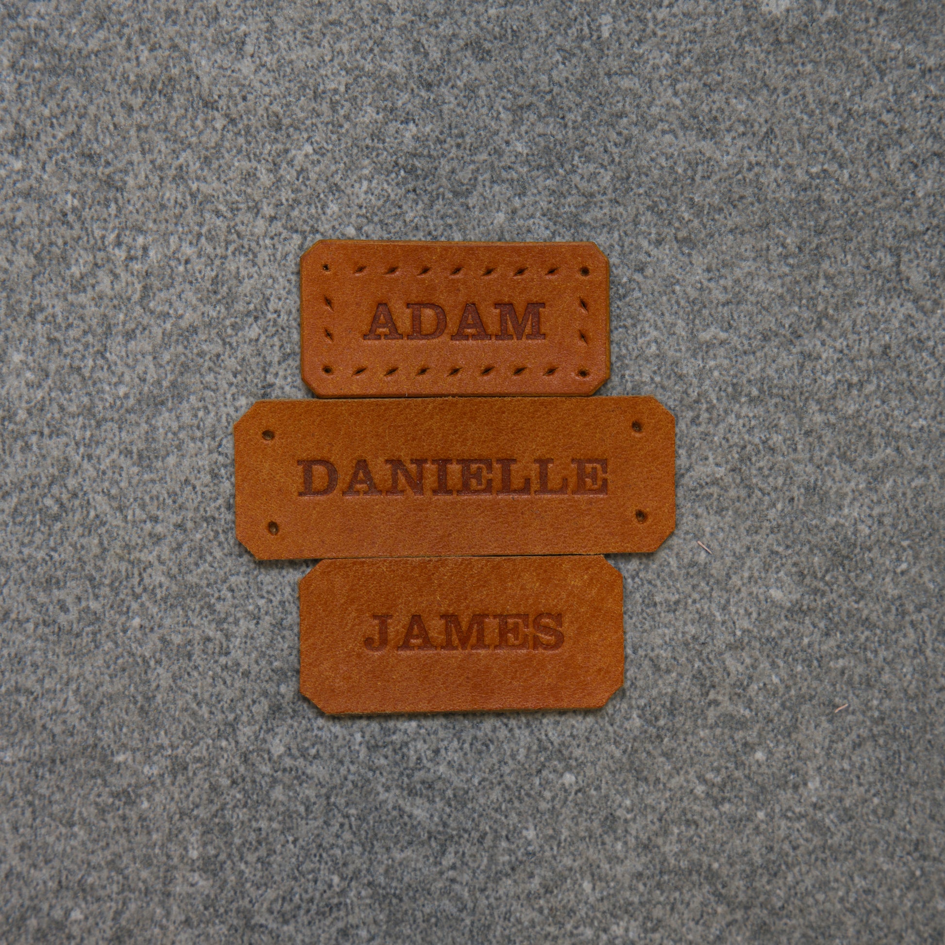 Three golden brown name tags labelled 'Adam', 'Danielle', and 'James' on a textured purple surface.