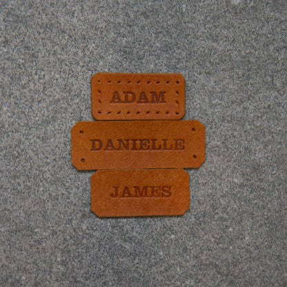 Three golden brown name tags labelled 'Adam', 'Danielle', and 'James' on a textured purple surface.