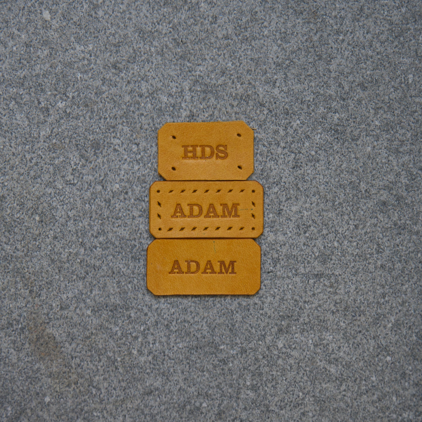 Three brown leather labels with 'HDS' and 'ADAM' text on a gray textured background