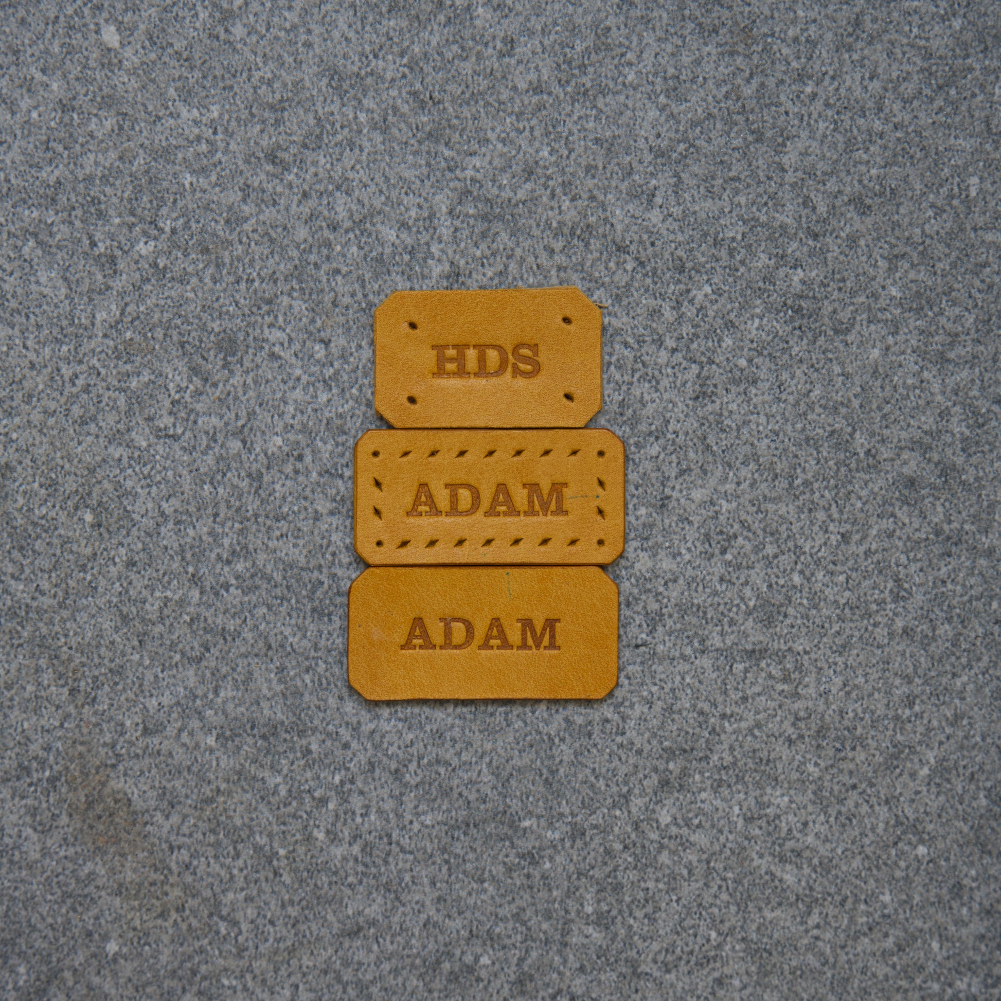 Three brown leather labels with 'HDS' and 'ADAM' text on a gray textured background