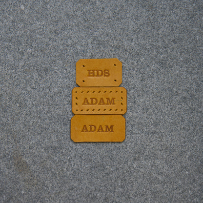 Three brown leather labels with 'HDS' and 'ADAM' text on a gray textured background