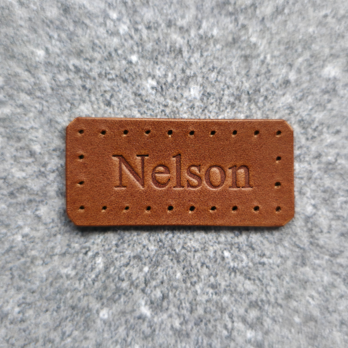 A rectangular brown leather patch with pre-drilled holes and the name 'Nelson' embossed in the center, shown on a textured gray background.