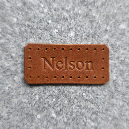 A rectangular brown leather patch with pre-drilled holes and the name 'Nelson' embossed in the center, shown on a textured gray background.