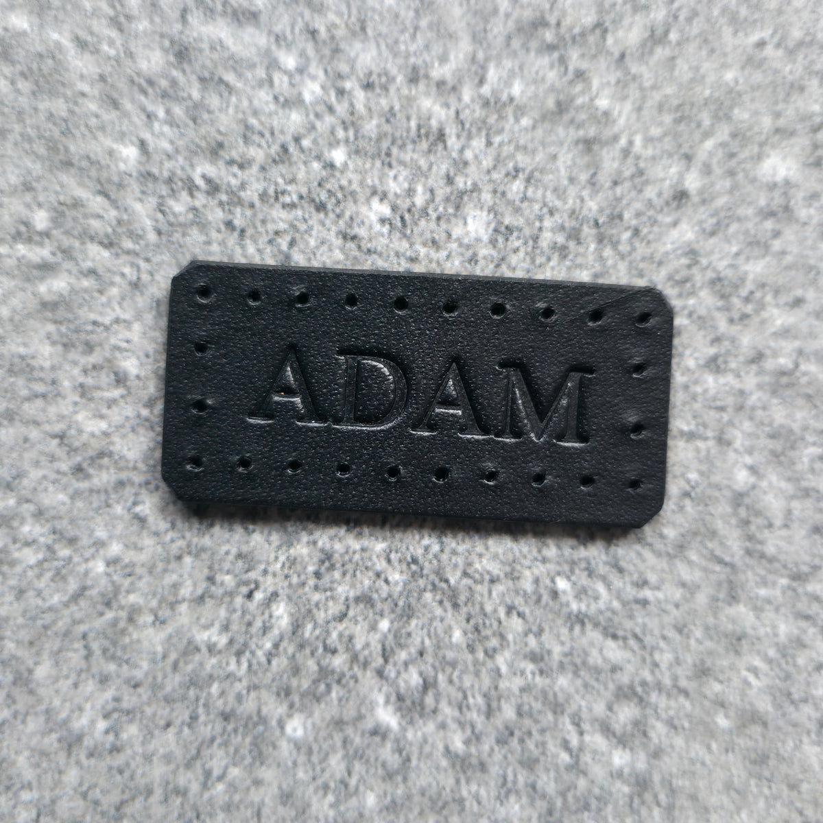 A rectangular black leather patch with the name 'ADAM' embossed in the center, surrounded by small perforations, placed on a textured gray fabric background.