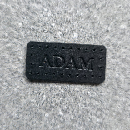 A rectangular black leather patch with the name 'ADAM' embossed in the center, surrounded by small perforations, placed on a textured gray fabric background.