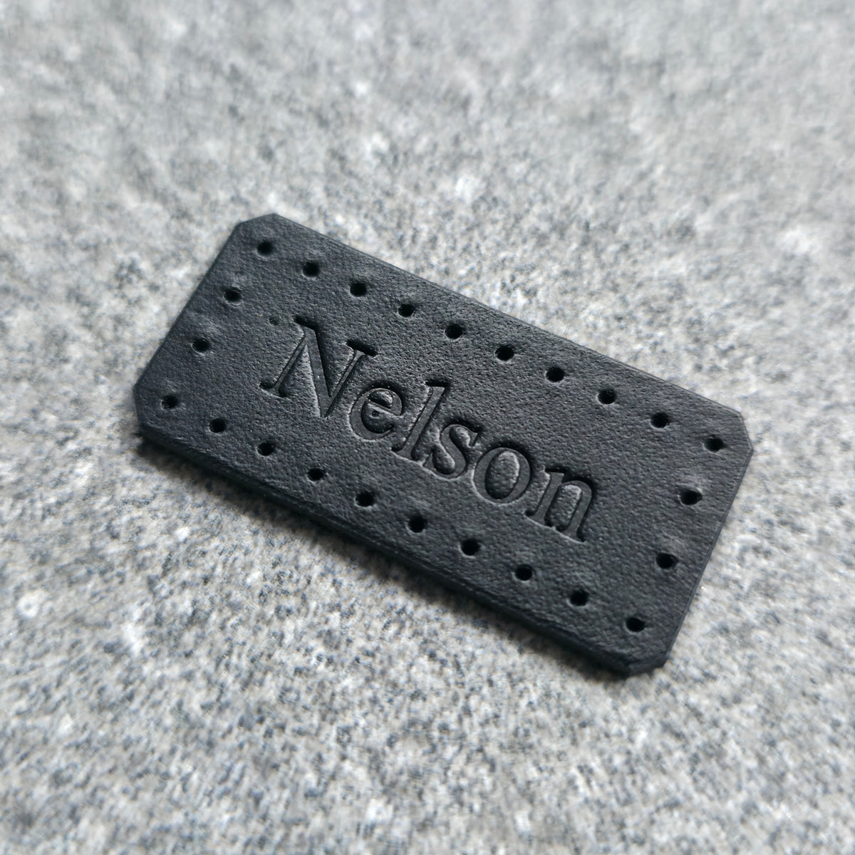 A black rectangular leather patch with pre-drilled holes and the name 'Nelson' embossed in the center, resting on a textured gray surface.