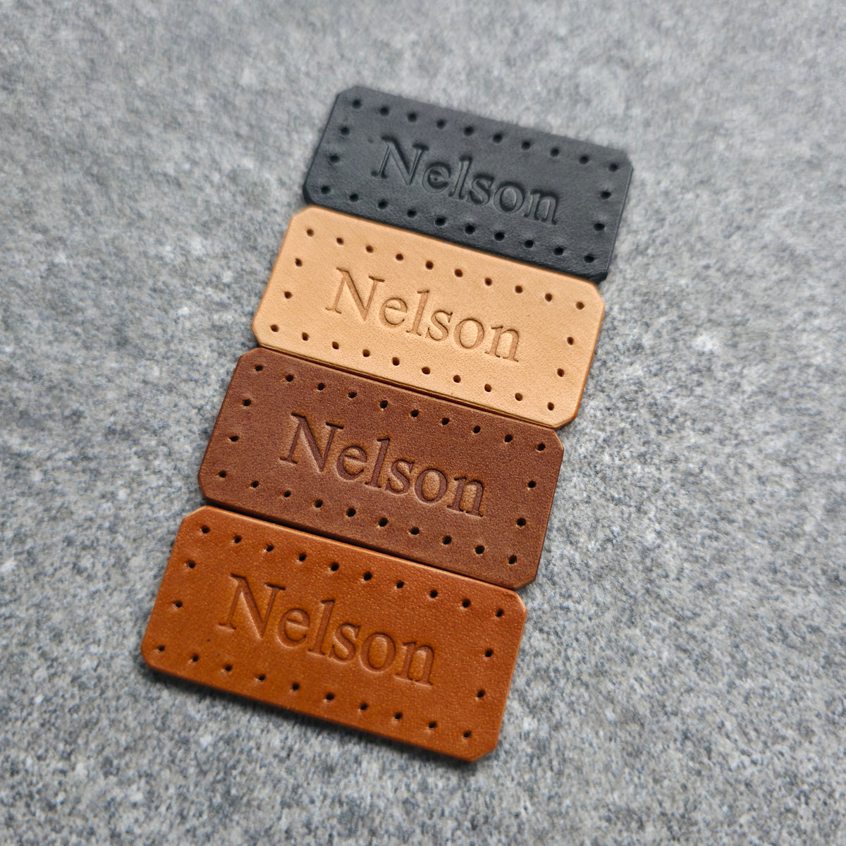 Four rectangular leather nameplates with perforated edges and the name 'Nelson' embossed, shown in black, tan, medium brown, and dark brown on a gray textured background.