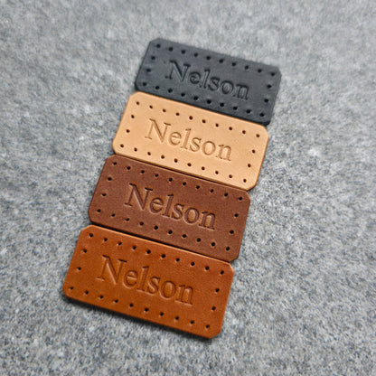 Four rectangular leather nameplates with perforated edges and the name 'Nelson' embossed, shown in black, tan, medium brown, and dark brown on a gray textured background.